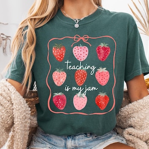 May include: Dark teal t-shirt featuring a strawberry design with the text "Teaching is my jam." The design includes nine strawberries in various shades of red and pink, framed by a pink border. The shirt is a casual, comfortable style.