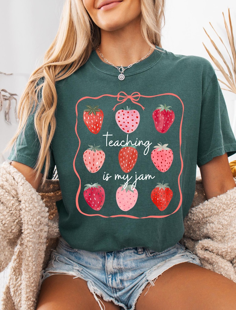 May include: Dark teal t-shirt featuring a strawberry design with the text "Teaching is my jam." The design includes nine strawberries in various shades of red and pink, framed by a pink border. The shirt is a casual, comfortable style.