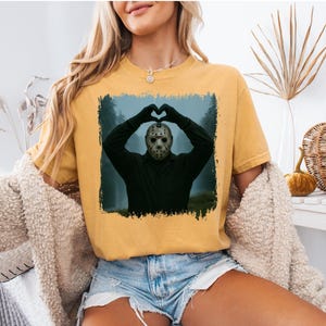以下が含まれることがあります： マスタードイエローのTシャツ。ホッケーマスクをかぶった人が手でハートを作っている額縁入りの画像が特徴です。画像は暗く霧のかかった背景です。Tシャツはデニムショーツと組み合わせています。