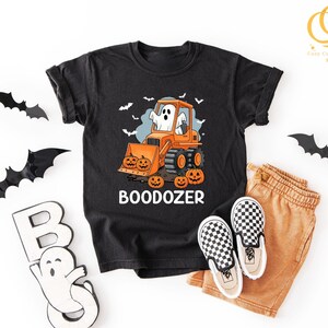 Peut inclure: T-shirt noir avec un motif d'Halloween repr&eacute;sentant un fant&ocirc;me conduisant un bulldozer orange avec des citrouilles. Le texte "BOODOZER" est en dessous. Accompagn&eacute; d'un short beige et de chaussures &agrave; enfiler &agrave; carreaux.
