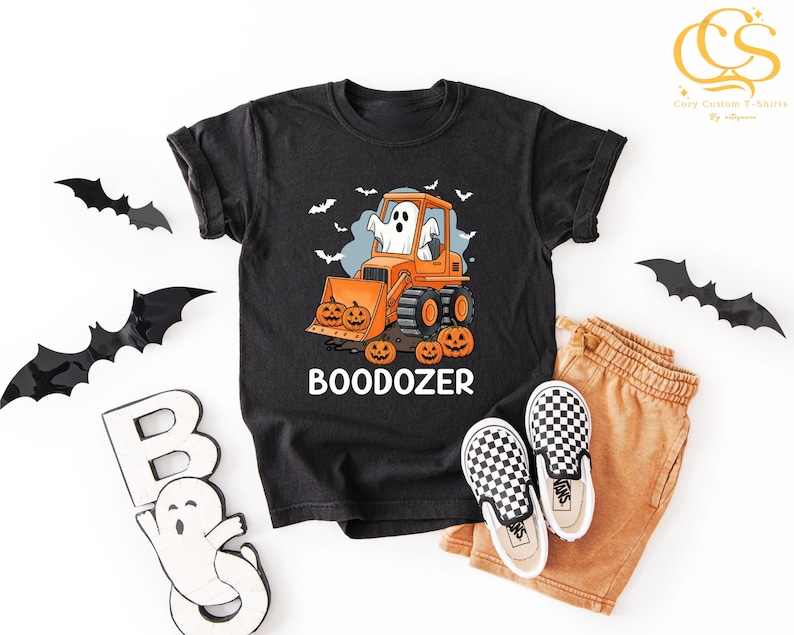 Peut inclure: T-shirt noir avec un motif d'Halloween repr&eacute;sentant un fant&ocirc;me conduisant un bulldozer orange avec des citrouilles. Le texte "BOODOZER" est en dessous. Accompagn&eacute; d'un short beige et de chaussures &agrave; enfiler &agrave; carreaux.