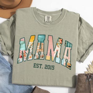 May include: Sage green t-shirt with the word "MAMA" in a patchwork design, featuring floral and plaid patterns. The text "EST. 2015" is below the word "MAMA". The shirt is folded, with a hat and jeans visible.