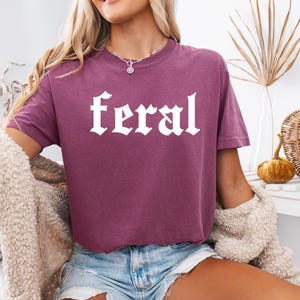 Pode incluir: Uma camiseta cropped malva com a palavra "feral" impressa em branco em uma fonte g&oacute;tica.