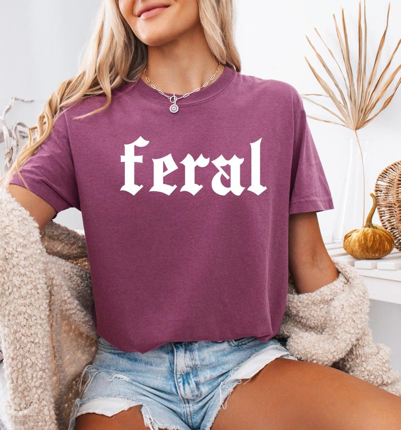 Pode incluir: Uma camiseta cropped malva com a palavra "feral" impressa em branco em uma fonte g&oacute;tica.