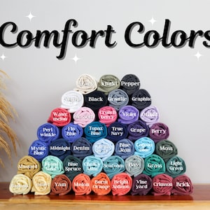 May include: A pyramid of rolled fabric swatches in various colors, labeled with names like "Khaki", "Pepper", "Black", "Granite", "Graphite", "Watermelon", "Blossom", "Berry", "Peri Winkle", "Flo Blue", "Topaz Blue", "True Navy", "Grape", "Berry", "Jean", "Mystic Blue", "Midnight", "Denim", "Blue Spruce", "Chalk Mint", "Seafoam", "Bay", "Grass", "Light Green", "Yam", "Melon", "Burnt Orange", "Bright Salmon", "Vine Yard", "Crimson", and "Brick". The text "Comfort Colors" is at the top of the image.