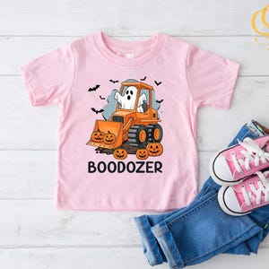 Peut inclure: T-shirt rose clair pour enfant avec un motif d'Halloween. On y voit un bulldozer orange conduit par un fant&ocirc;me, des citrouilles et le mot "BOODOZER". Des chauves-souris noires volent en arri&egrave;re-plan. Le tout est accompagn&eacute; d'un jean bleu et de chaussures roses.