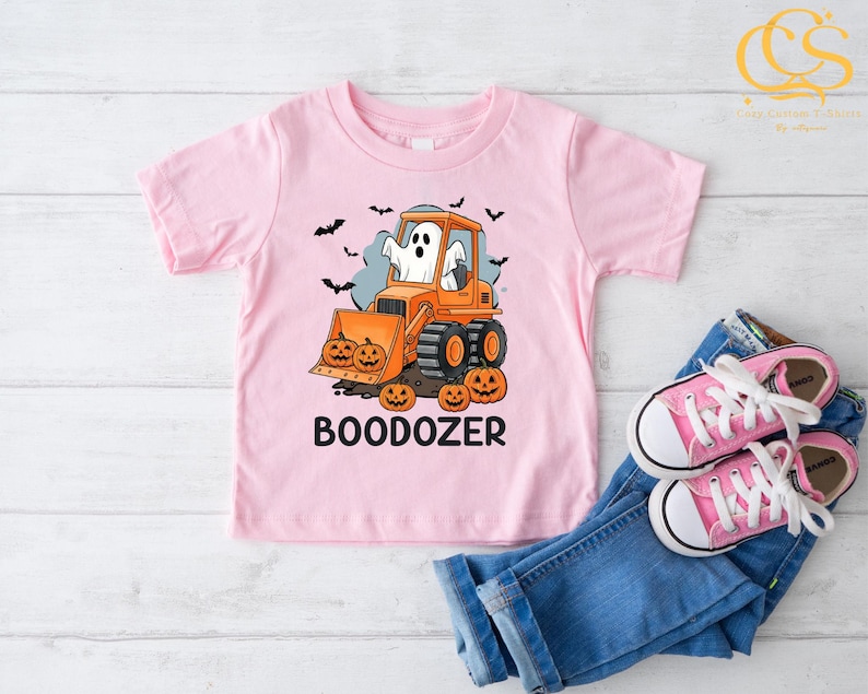 Peut inclure: T-shirt rose clair pour enfant avec un motif d'Halloween. On y voit un bulldozer orange conduit par un fant&ocirc;me, des citrouilles et le mot "BOODOZER". Des chauves-souris noires volent en arri&egrave;re-plan. Le tout est accompagn&eacute; d'un jean bleu et de chaussures roses.