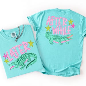 May include: Two turquoise t-shirts with a graphic of a green alligator and the text "LATER" and "AFTER WHILE" in pink. The shirts also feature pink and yellow star graphics. A silver chain necklace is on one shirt.