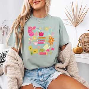 May include: A light blue t-shirt with a colorful graphic design featuring school supplies, flowers, and an apple. The graphic includes a pencil, a ruler, a book, a paperclip, a flower, and an apple.