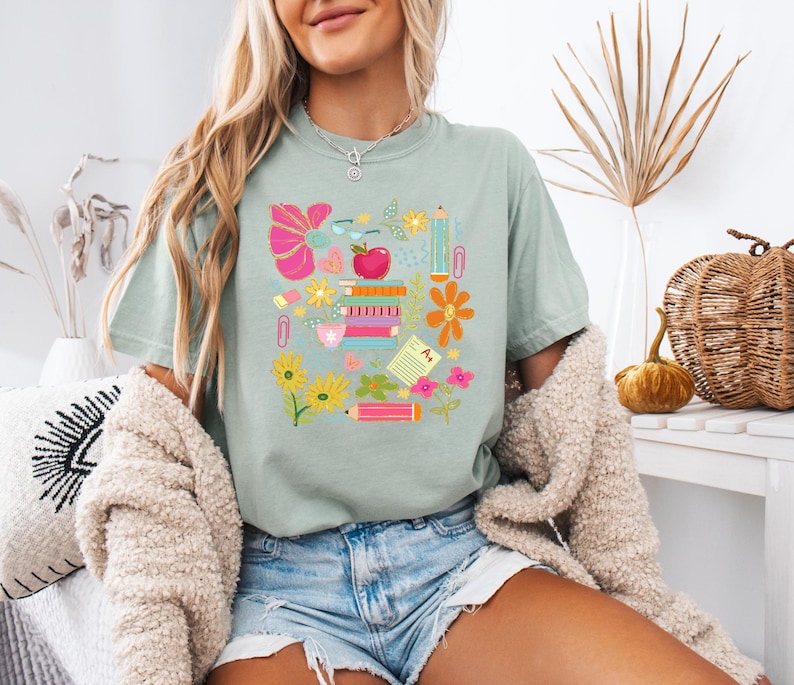 May include: A light blue t-shirt with a colorful graphic design featuring school supplies, flowers, and an apple. The graphic includes a pencil, a ruler, a book, a paperclip, a flower, and an apple.
