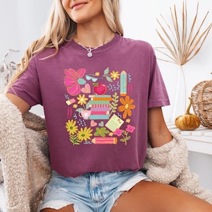 May include: A purple cropped t-shirt with a colorful graphic design featuring school supplies, flowers, and an apple. The design includes a stack of books, pencils, a ruler, a cup of coffee, and a flower.