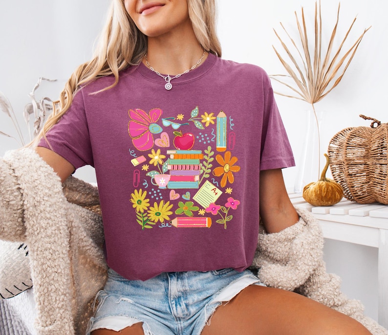 May include: A purple cropped t-shirt with a colorful graphic design featuring school supplies, flowers, and an apple. The design includes a stack of books, pencils, a ruler, a cup of coffee, and a flower.