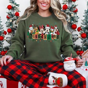 K&ouml;nnte beinhalten: Ein olivgr&uuml;nes Sweatshirt mit dem Schriftzug "Disneyland" und Disney-Figuren mit Weihnachtsm&uuml;tzen. Die Person tr&auml;gt eine rot-schwarz karierte Pyjamahose und h&auml;lt einen Weihnachtsmann-Becher.