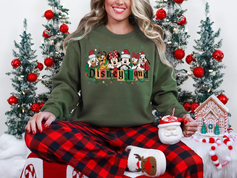 K&ouml;nnte beinhalten: Ein olivgr&uuml;nes Sweatshirt mit dem Schriftzug "Disneyland" und Disney-Figuren mit Weihnachtsm&uuml;tzen. Die Person tr&auml;gt eine rot-schwarz karierte Pyjamahose und h&auml;lt einen Weihnachtsmann-Becher.