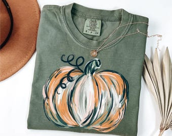 May include: Olive green t-shirt with a painted pumpkin design in orange, white, and green. The shirt has a crew neck and short sleeves. A gold necklace with a small pendant is draped across the shirt. A brown hat is in the background.