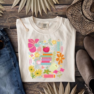 May include: A white t-shirt with a colorful graphic design featuring a stack of books, pencils, flowers, and other school supplies. The design is surrounded by a border of pink, orange, and yellow flowers.