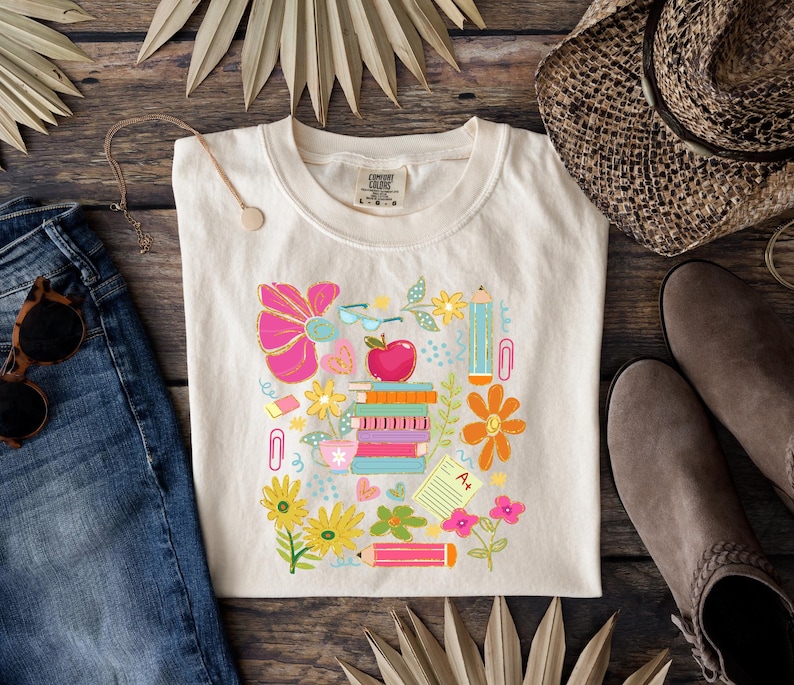 May include: A white t-shirt with a colorful graphic design featuring a stack of books, pencils, flowers, and other school supplies. The design is surrounded by a border of pink, orange, and yellow flowers.