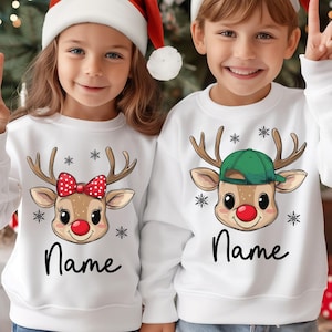 Peut inclure: Deux enfants portant des pulls de Noël blancs avec des motifs de rennes et le mot "Name" en dessous. Un renne a un nœud rouge, l'autre une casquette verte. Les deux enfants portent des bonnets de Père Noël et font le signe de la victoire.