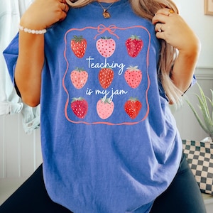 May include: Blue t-shirt with a strawberry design and the text "Teaching is my jam." The design features nine strawberries in various shades of red and pink, framed by a pink border with a bow at the top. The shirt is a comfortable, casual style.