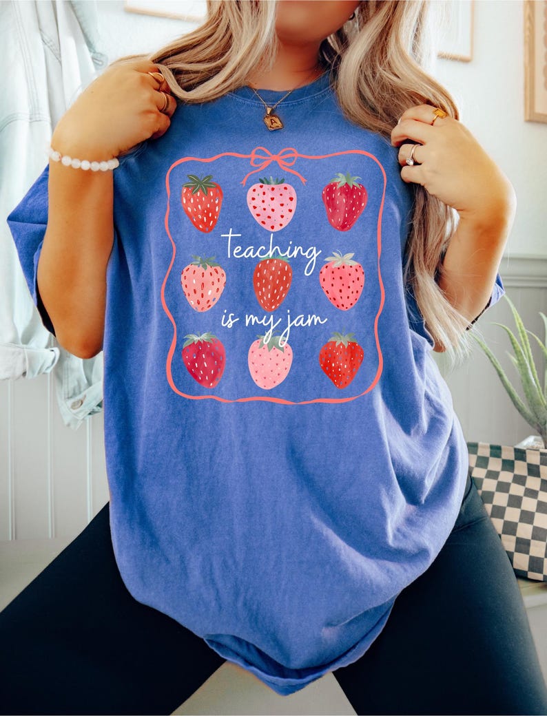 May include: Blue t-shirt with a strawberry design and the text "Teaching is my jam." The design features nine strawberries in various shades of red and pink, framed by a pink border with a bow at the top. The shirt is a comfortable, casual style.