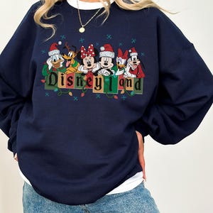 K&ouml;nnte beinhalten: Marineblaues Sweatshirt mit Disney-Figuren, die Weihnachtsm&uuml;tzen tragen. Das Design umfasst Donald Duck, Pluto, Minnie Maus, Mickey Maus und Goofy, mit dem Text "Disneyland" darunter. Das Sweatshirt hat einen wei&szlig;en Kragen.