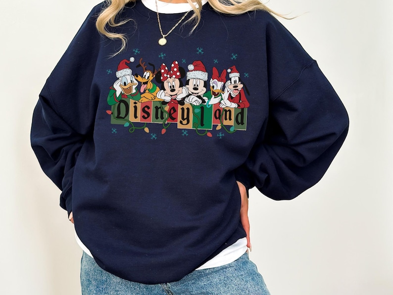 K&ouml;nnte beinhalten: Marineblaues Sweatshirt mit Disney-Figuren, die Weihnachtsm&uuml;tzen tragen. Das Design umfasst Donald Duck, Pluto, Minnie Maus, Mickey Maus und Goofy, mit dem Text "Disneyland" darunter. Das Sweatshirt hat einen wei&szlig;en Kragen.