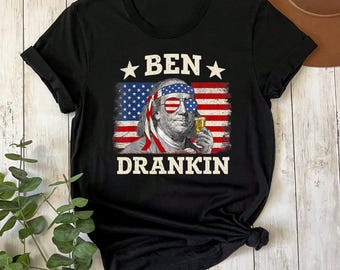 May include: Black t-shirt with a graphic of Benjamin Franklin wearing sunglasses and a headband with the American flag design. The text "BEN DRANKIN" is printed above and below the image in white.