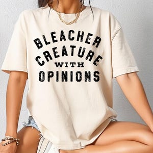 May include: Cream-colored t-shirt with the text "BLEACHER CREATURE WITH OPINIONS" in distressed black lettering. The shirt has short sleeves and a relaxed fit, suitable for casual wear.