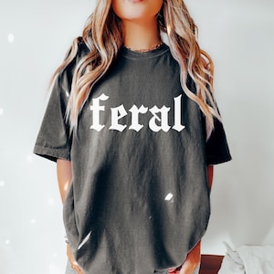Pode incluir: Uma camiseta cinza escura de tamanho grande com a palavra "feral" impressa em branco em uma fonte g&oacute;tica.