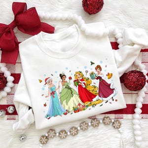 Puede incluir: Sudadera blanca de cuello redondo con un gráfico festivo de cuatro princesas de dibujos animados. Las princesas visten trajes navideños y están rodeadas de copos de nieve y regalos. La sudadera está decorada con adornos navideños y un lazo rojo.
