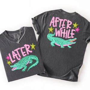 May include: Two dark gray t-shirts with a vintage wash. One shirt features the text "LATER" and a green alligator graphic. The other shirt has "AFTER WHILE" and the same alligator design. Both designs include pink and yellow accents.