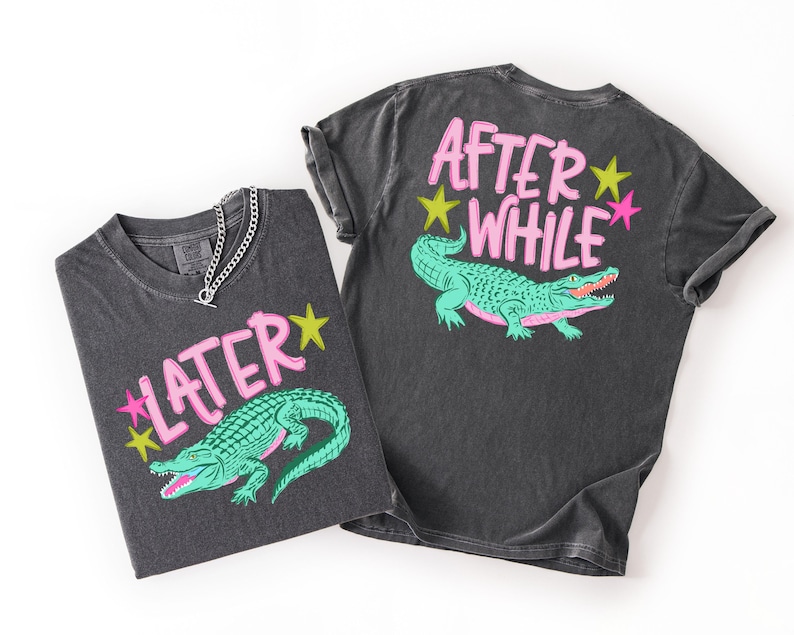 May include: Two dark gray t-shirts with a vintage wash. One shirt features the text "LATER" and a green alligator graphic. The other shirt has "AFTER WHILE" and the same alligator design. Both designs include pink and yellow accents.