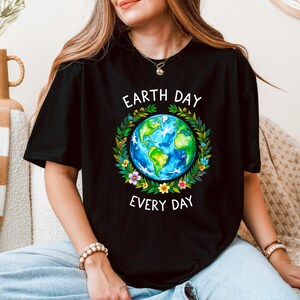 Puede incluir: Una camiseta negra con un gr&aacute;fico de una Tierra colorida rodeada de flores y hojas. El texto "Earth Day Every Day" est&aacute; impreso en blanco debajo del gr&aacute;fico.