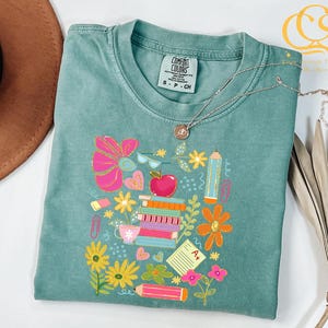 May include: A teal blue t-shirt with a colorful graphic design of school supplies, including books, pencils, an apple, and flowers. The text "COMFORT COLORS" and "S-P-CH" are visible on the shirt.