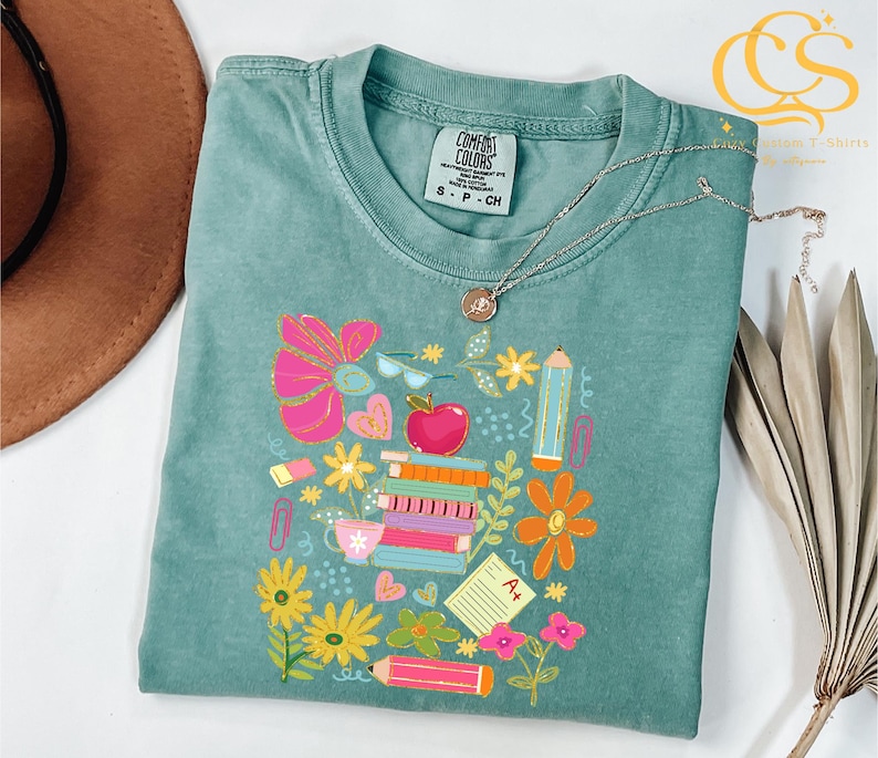 May include: A teal blue t-shirt with a colorful graphic design of school supplies, including books, pencils, an apple, and flowers. The text "COMFORT COLORS" and "S-P-CH" are visible on the shirt.