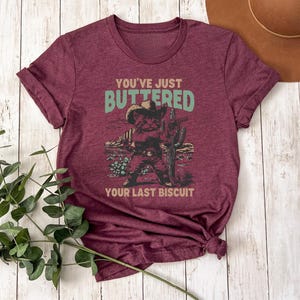 May include: Burgundy t-shirt with the text "You've Just Buttered Your Last Biscuit" above a graphic of a cat in a cowboy hat and boots, holding a cactus. The design is in shades of green, brown, and tan.