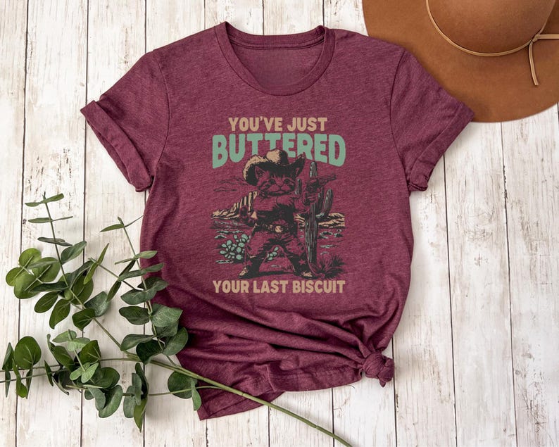 May include: Burgundy t-shirt with the text "You've Just Buttered Your Last Biscuit" above a graphic of a cat in a cowboy hat and boots, holding a cactus. The design is in shades of green, brown, and tan.