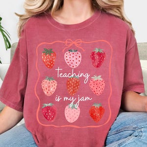 May include: A maroon-colored t-shirt featuring a strawberry design. The design includes nine strawberries in various shades of red and pink, arranged within a pink frame. The text "Teaching is my jam" is written in cursive.