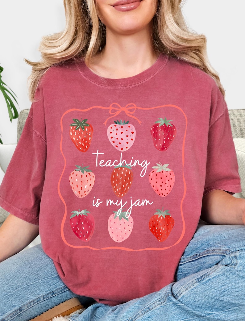 May include: A maroon-colored t-shirt featuring a strawberry design. The design includes nine strawberries in various shades of red and pink, arranged within a pink frame. The text "Teaching is my jam" is written in cursive.