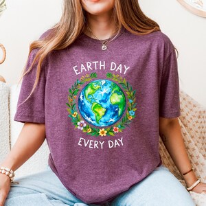 Puede incluir: Una camiseta morada con un gr&aacute;fico de la Tierra rodeada de flores y el texto "Earth Day Every Day".