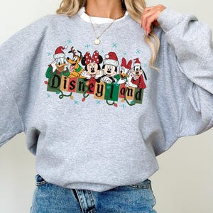 K&ouml;nnte beinhalten: Ein hellgraues Sweatshirt mit einer farbenfrohen Grafik von Disney-Figuren mit Weihnachtsm&uuml;tzen. Das Wort "Disneyland" wird in einer festlichen Schriftart mit Lichterketten dargestellt. Ein gem&uuml;tliches Oberteil mit langen &Auml;rmeln f&uuml;r Disney-Fans.