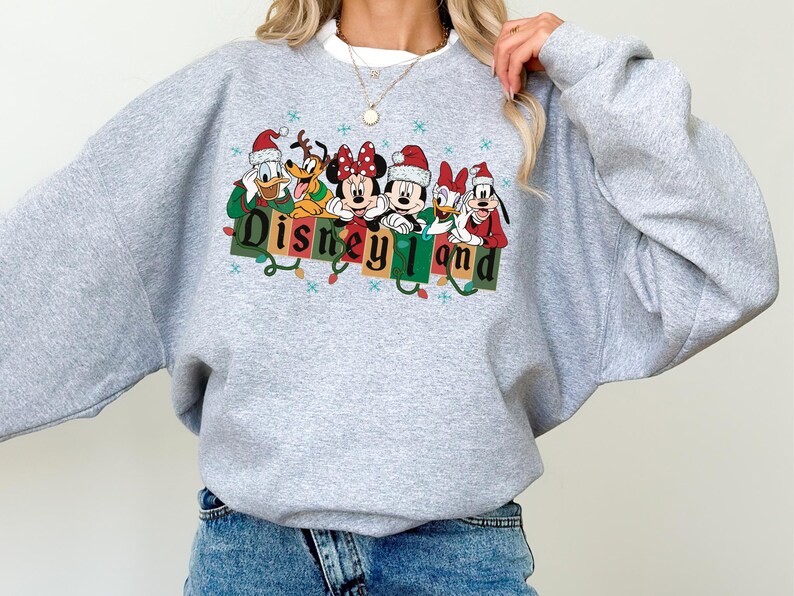 K&ouml;nnte beinhalten: Ein hellgraues Sweatshirt mit einer farbenfrohen Grafik von Disney-Figuren mit Weihnachtsm&uuml;tzen. Das Wort "Disneyland" wird in einer festlichen Schriftart mit Lichterketten dargestellt. Ein gem&uuml;tliches Oberteil mit langen &Auml;rmeln f&uuml;r Disney-Fans.