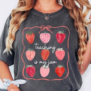 May include: Dark gray t-shirt featuring a strawberry design with the text "Teaching is my jam." The design includes nine strawberries in various shades of red and pink, framed by a pink border with a bow at the top. The shirt is a casual wear item.
