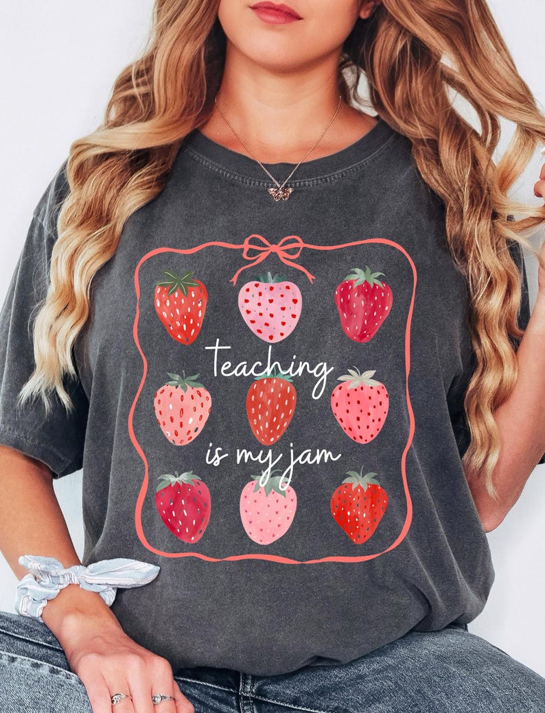 May include: Dark gray t-shirt featuring a strawberry design with the text "Teaching is my jam." The design includes nine strawberries in various shades of red and pink, framed by a pink border with a bow at the top. The shirt is a casual wear item.