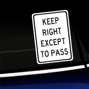 Può includere: Un adesivo rettangolare bianco con bordo nero e la scritta "KEEP RIGHT EXCEPT TO PASS" in grassetto nero. L'adesivo è attaccato a una superficie scura, probabilmente un finestrino dell'auto.