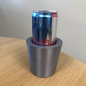 May include: A gray 3D printed can holder with a silver finish. A blue and red Red Bull energy drink can is inside the holder.