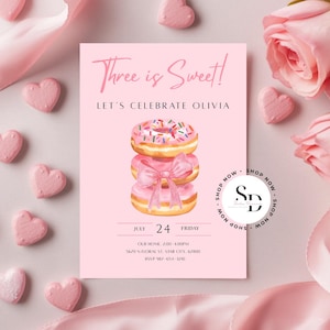 Può includere: Un biglietto d'invito rosa con il testo "Three is Sweet!" e "Let's Celebrate Olivia." Presenta una pila di ciambelle ad acquerello con un fiocco rosa. Caramelle rosa a forma di cuore e rose circondano la carta.