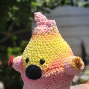 Könnte beinhalten: Handgefertigtes Häkeltier in einem skurrilen Design. Das Spielzeug hat einen Farbverlauf aus Rosa, Gelb und Orange, mit schwarzen Knopfaugen und einer dunkelblauen Nase. Es hat kleine Arme und Füße und eine strukturierte Oberfläche.