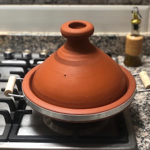 May include: A terracotta tagine pot with a conical lid and a metal base, on a gas hob. The pot has wooden handles and is a warm, earthy orange colour. An olive oil dispenser is in the background.