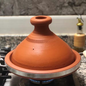 Può includere: Una pentola tagine in terracotta con coperchio conico e bordo argentato, appoggiata su un fornello a gas. La pentola è di un caldo colore marrone-arancio e il coperchio presenta linee incise decorative. Il bruciatore del fornello è acceso con una fiamma blu.